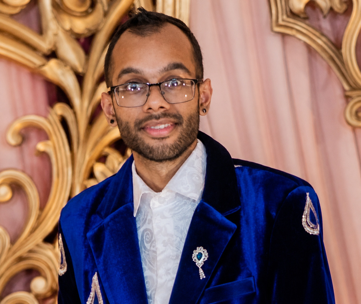 Sahir Maharaj smiling in glasses and a deep blue embroidered jacket, photographed against a soft pink backdrop